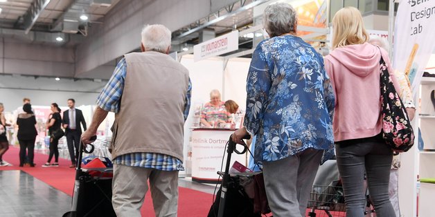 Ein älterer Mann und zwei Frauen mit Rollatoren - nur von hinten zu sehen -  laufen durch die Messehallen auf dem Deutschen Seniorentag in Dortmund.