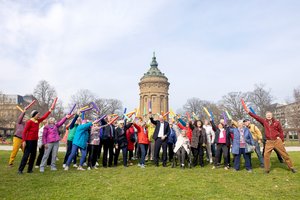 Eine Gruppe älterer Menschen machen in einem Park gemeinsam Sport. Im Hintergrund sieht man einen alten Turm. Sie haben kurze, bunte Schwimmnudeln in der Hand. Sie strecken sie nach vorne und in die Höhe.