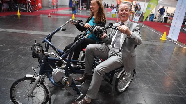 Franz Müntefering fährt mit einer jungen Frau auf einem Dreirad über die Messe auf dem Deutschen Seniorentag und lacht in die Kamera.