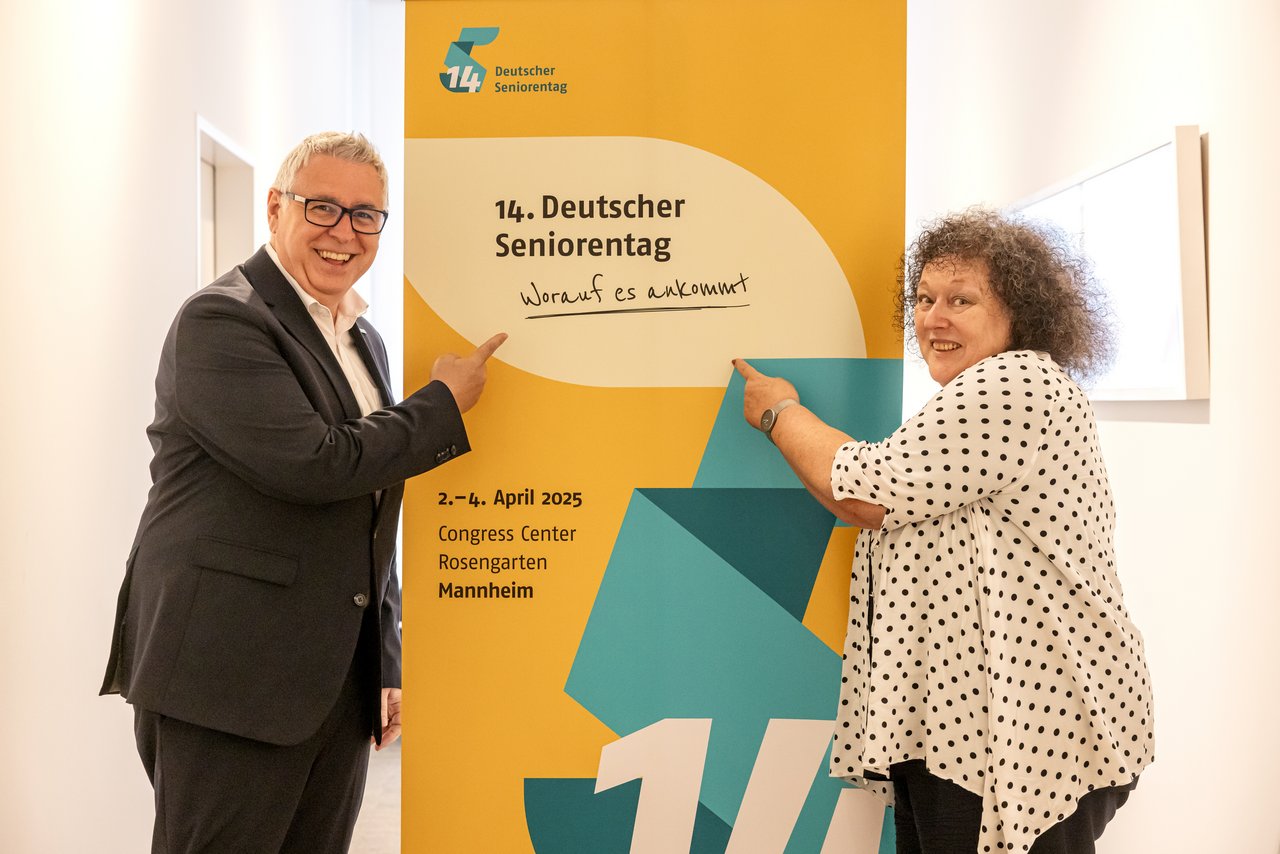 Eine ältere Frau und ein älterer Mann zeigen lächelnd auf das Aufstellbanner des 14. Deutschen Seniorentages. Klick öffnet eine vergrößerte Ansicht.