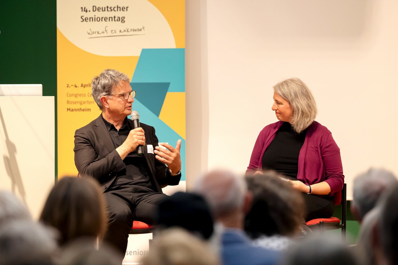 Ein Mann sitzt mit Mikrofon auf einem Stuhl vor dem Aufstellbanner des 14. Deutschen Seniorentages und spicht zu einer Frau, welche ihm gegenüber sitzt. Klick öffnet eine vergrößerte Ansicht.
