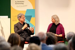 Ein Mann sitzt mit Mikrofon auf einem Stuhl vor dem Aufstellbanner des 14. Deutschen Seniorentages und spicht zu einer Frau, welche ihm gegenüber sitzt.
